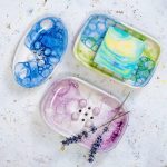 Ročno izdelan ovalni keramični milnik iz bele gline, dekoriran s tehniko mehurčkanja v svežih modrih, zelenih in lila tonih. Gumbi in odtočne luknjice ohranjajo milo suho, mehurčkast vzorec pa doda kopalnici igriv, peneč značaj.