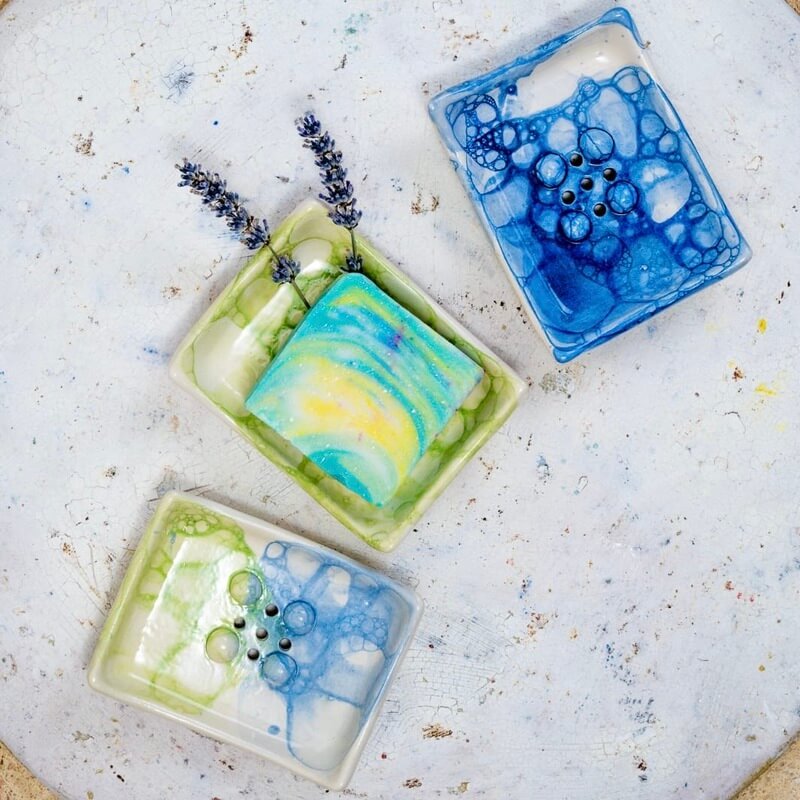 Ročno izdelan pravokotni keramični milnik iz bele gline, dekoriran s tehniko mehurčkanja v svežih modrih in zelenih tonih. Gumbi in odtočne luknjice ohranjajo milo suho, mehurčkast vzorec pa doda kopalnici igriv, peneč značaj.